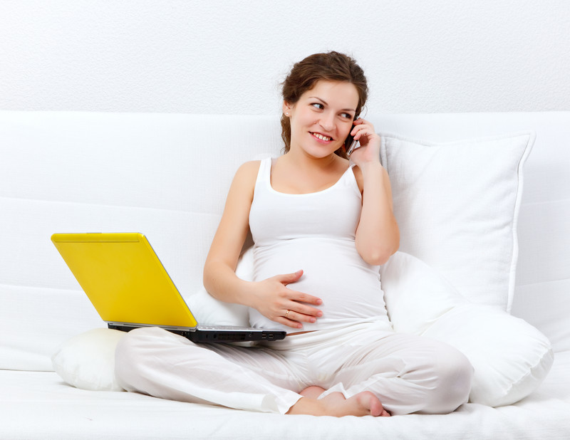 Schwangere Frau telefoniert, auf dem Bett ein Laptop