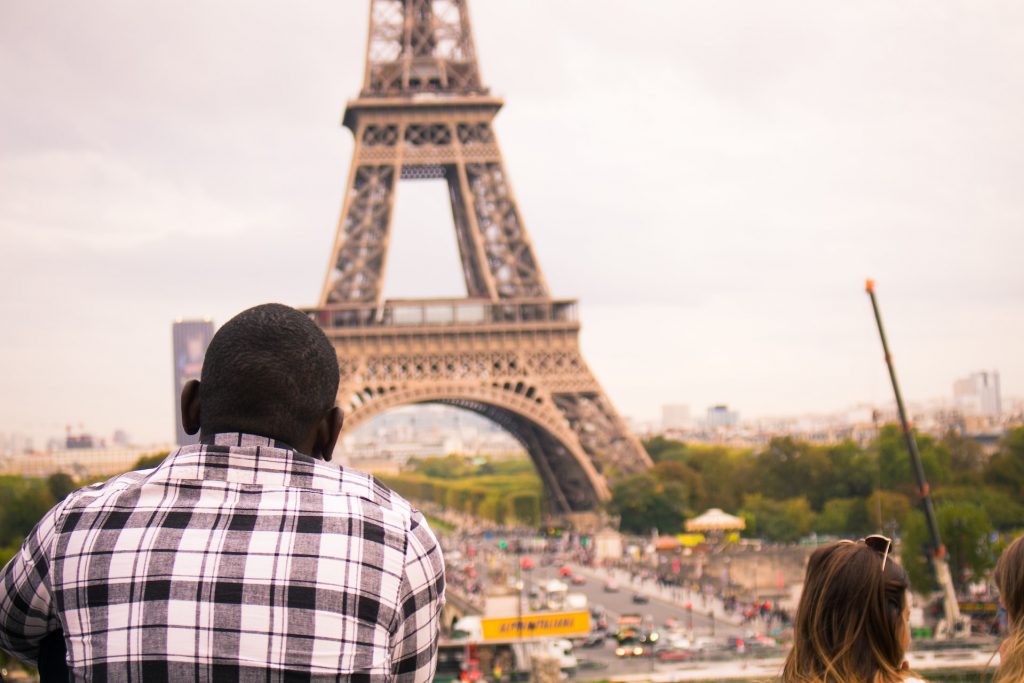 Ausblick auf den Eiffelturm in Paris, erlebt durch die Augen eines Touristen. Ein Mann und eine Frau bestaunen das berühmte Wahrzeichen, während im Hintergrund die geschäftige Stadt Paris zu sehen ist.